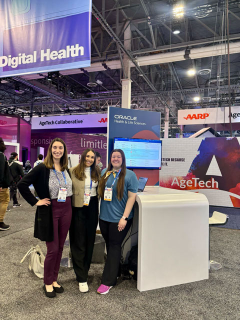 Three women wearing conference badges stand smiling at an Oracle digital health exhibition booth. They pose beside a white kiosk with a screen displaying Oracle Health & Life Sciences software. Overhead signage reads “Digital Health,” with surrounding banners referencing AgeTech and healthcare innovation in a busy convention hall. 