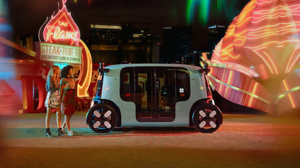 Two people, dressed in stylish evening wear, stand next to a Zoox autonomous robotaxi on a city street at night. Bright, neon signs from a diner or casino are visible in the background. 
