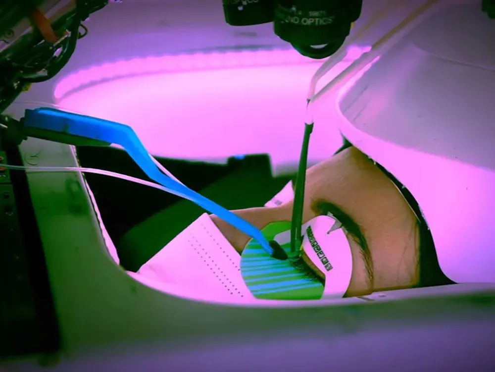 A close-up shot of a person's eye receiving eyelash extensions from a small robotic arm (LUUM Precision Lash). The area is lit with a pink/purple light, and a blue and white applicator is visible near the person's eye.